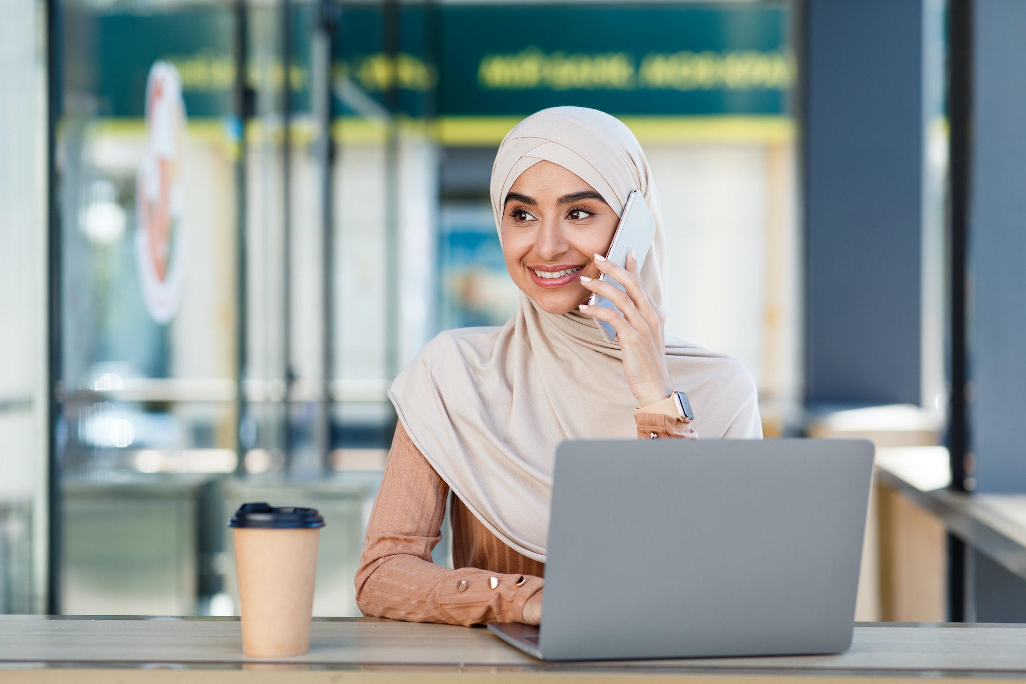 Smiling friendly cute millennial arab muslim woman in hijab speaks on phone and types on laptop in